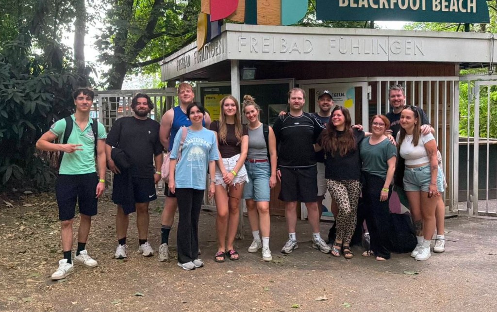 Das REVOIC Team bei einem Ausflug zum Blackfoot Beach im Freibad Fühlingen. 11 Mitarbeitende stehen lächelnd vor dem Eingangsbereich.